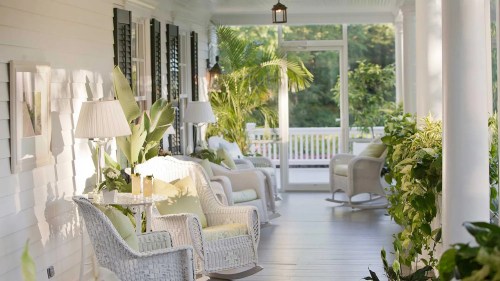 A serene porch with white wicker chairs, lamps, and lush greenery, creating a relaxing outdoor space.
