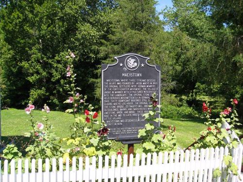 Historical marker for Maestown surrounded by flowers and a white picket fence, set against a green landscape.