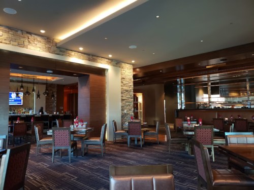 Modern restaurant interior with wooden tables, chairs, and a bar area, featuring stone accents and soft lighting.