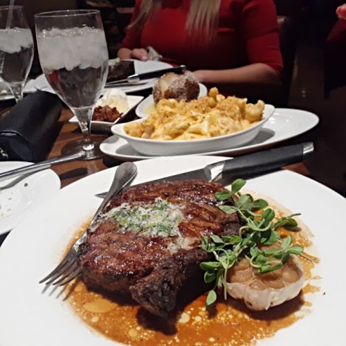 A plate with a grilled steak, garlic, and herbs, accompanied by macaroni and cheese and drinks in the background.