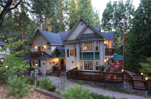 A charming two-story house surrounded by trees, featuring a wraparound deck and warm outdoor lighting.