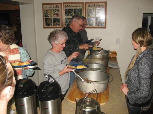 A group of people serving themselves food from large pots at a communal dining area.