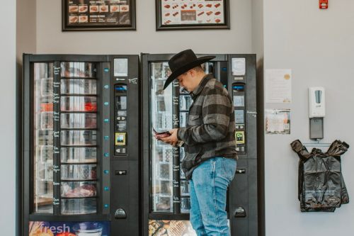 A man in a cowboy hat and plaid shirt stands by vending machines, checking his phone in a casual setting.