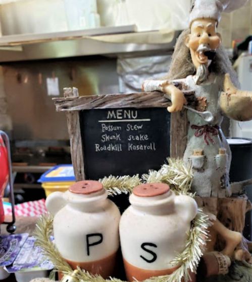 A whimsical chef figure beside a menu listing unusual dishes like possum stew and skunk steak, with decorative jars below.