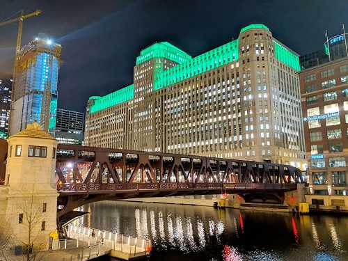 A night view of a bridge over a river, with a brightly lit green building in the background.