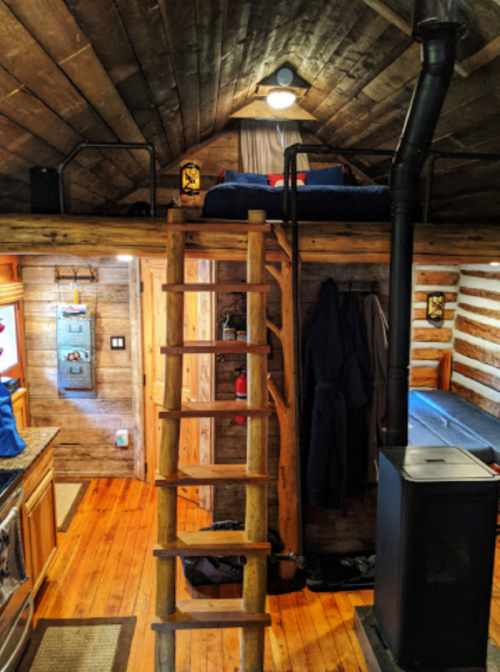 Cozy interior of a log cabin with a loft bed, wooden ladder, and rustic kitchen features. Warm, inviting atmosphere.