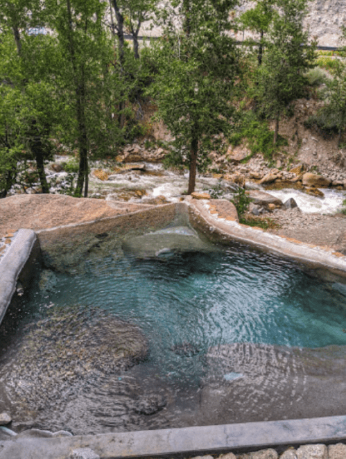 A natural hot spring surrounded by trees, with a flowing river in the background and rocky terrain.