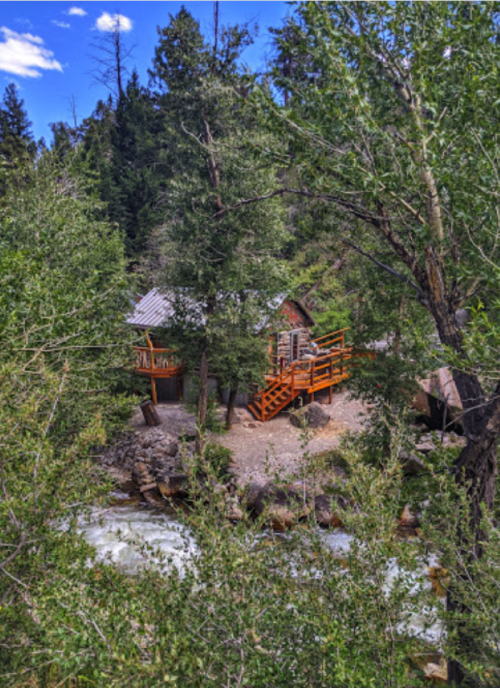 A cozy cabin surrounded by trees, with a wooden deck overlooking a flowing river. Bright blue sky above.