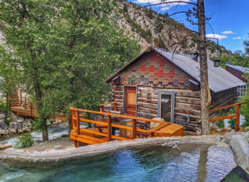 A cozy log cabin with a wooden deck beside a stream, surrounded by trees and mountains. A hot tub is in the foreground.