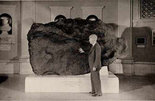 A man stands beside a large meteorite displayed in a museum, highlighting its size and significance.