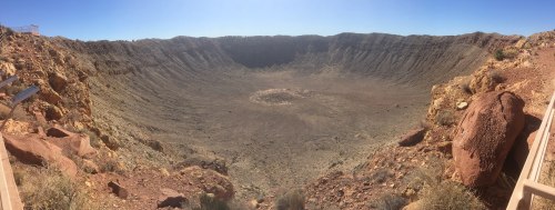 A panoramic view of a large, dry crater surrounded by rocky terrain under a clear blue sky.