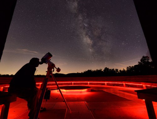 A person observes the night sky through a telescope, surrounded by red lighting and a backdrop of stars and the Milky Way.