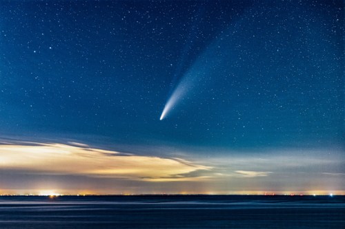 A comet streaks across a starry night sky above a calm, dark landscape with distant lights on the horizon.
