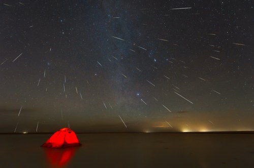A red tent sits on a calm shore under a starry sky filled with shooting stars and the Milky Way.