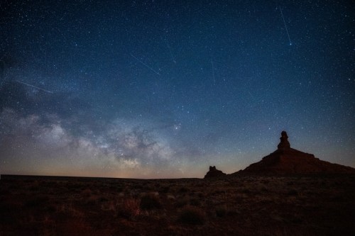 A starry night sky with the Milky Way and shooting stars above a rocky landscape.
