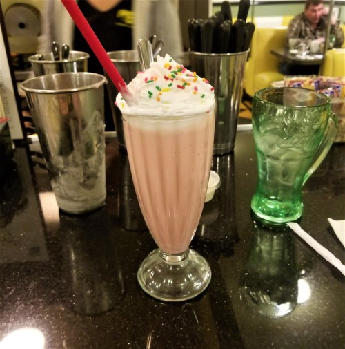 A pink milkshake topped with whipped cream and sprinkles, served in a tall glass on a black table.