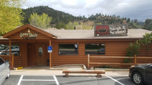 A log cabin-style restaurant with a sign reading "Steak & Seafood," surrounded by trees and mountains.