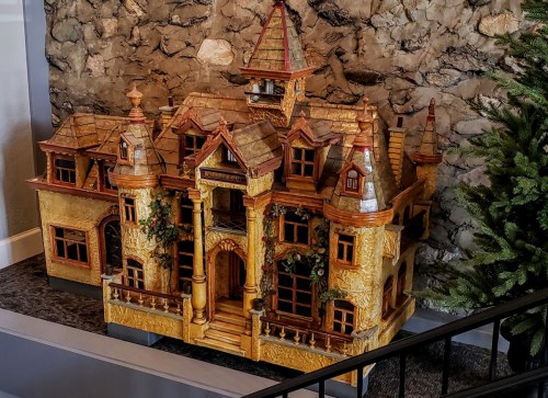 A detailed wooden dollhouse with multiple stories, intricate architecture, and decorative elements, set against a stone wall.