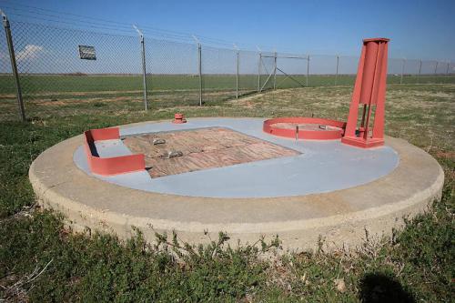 A circular concrete platform with a metal cover and a red structure, set in an open field with a fence in the background.