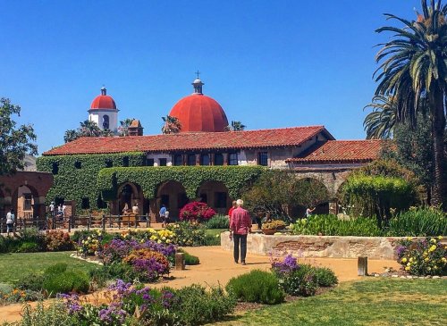 A person walks through a vibrant garden with a historic building featuring red domes and lush greenery.