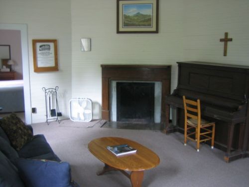 Cozy living room with a piano, a wooden coffee table, a fan, and a fireplace, featuring a mountain painting on the wall.