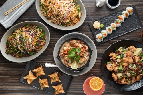 A spread of Asian dishes including noodles, sushi, shrimp, and dumplings on a wooden table.