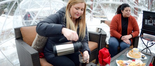 A woman pours a drink while another woman sits nearby, both enjoying a cozy setting in a snow-covered igloo.