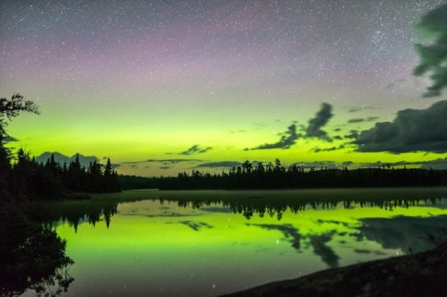 A serene lake reflects a vibrant green aurora under a starry sky, surrounded by dark silhouettes of trees.