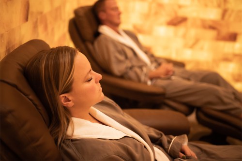 Two people in spa robes relax in reclining chairs, surrounded by warm, glowing salt wall decor.