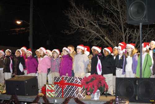 A group of children wearing Santa hats sings on stage, surrounded by holiday decorations and gifts.