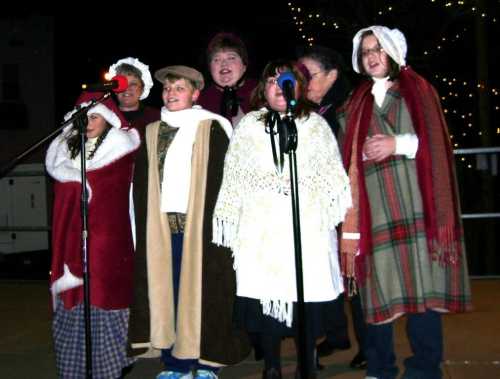 A group of children and adults in festive attire singing into microphones at an outdoor event.