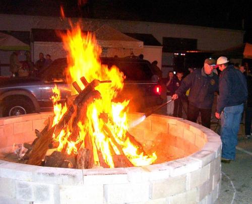 A large bonfire burns in a stone pit, with people gathered around in the background at night.
