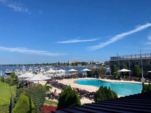 A sunny view of a pool area with lounge chairs, surrounded by greenery and boats in the water under a clear blue sky.