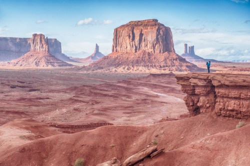 A lone hiker stands on a rocky ledge overlooking a vast desert landscape with towering red rock formations.