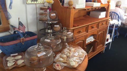 A wooden table displays various baked goods under glass cloches, with a blue basket and a person seated in the background.