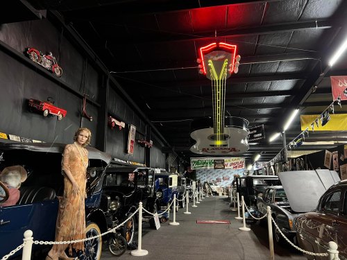 A vintage car museum aisle featuring classic cars, a neon sign, and a mannequin in 1920s attire.
