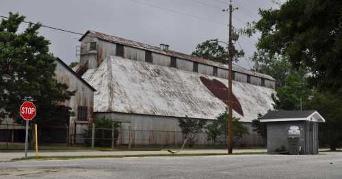 Old, weathered barn with a rusty roof, near a stop sign and a small building, under a cloudy sky.