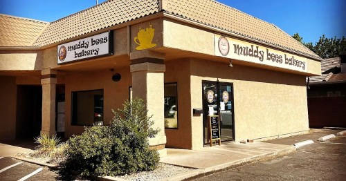Exterior of Muddy Bees Bakery, featuring a tan building with a sign and a small entrance, surrounded by greenery.