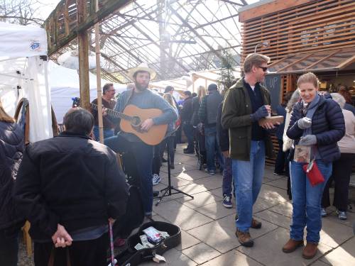 A lively market scene with a musician playing guitar, people browsing, and enjoying food in a greenhouse setting.