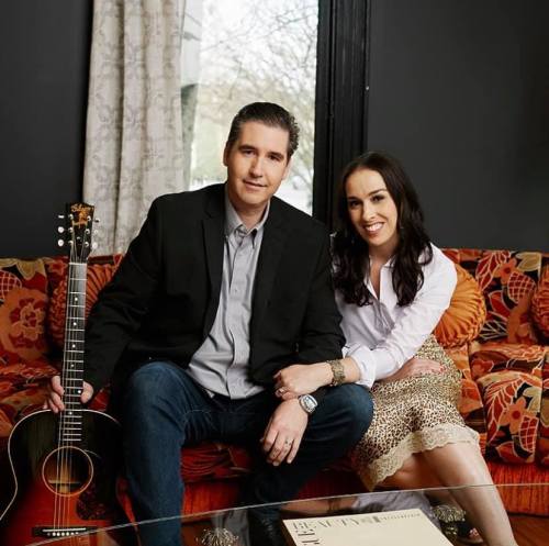 A man and woman sit together on a colorful couch, the man holding an acoustic guitar. They smile at the camera.