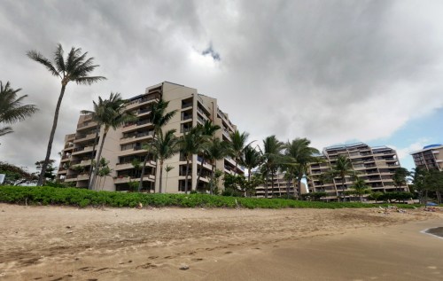 Beachfront view of modern apartment buildings surrounded by palm trees under a cloudy sky.