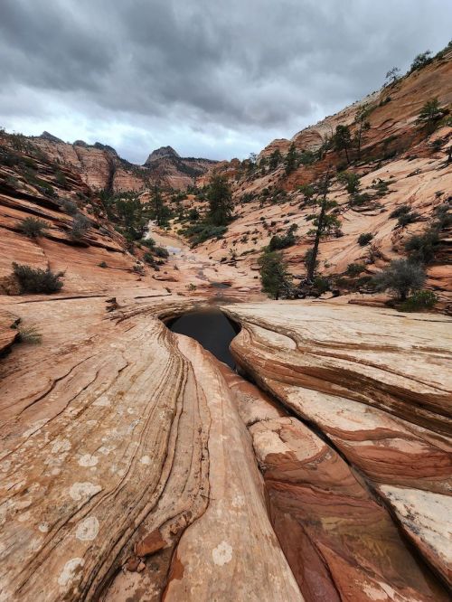 A rocky canyon landscape with layered red and tan rock formations under a cloudy sky. A small pool of water is visible.