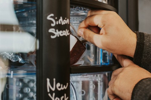 A hand reaches for a package labeled "Sidon Steak" in a refrigerator with various food items.