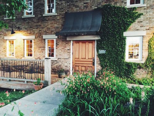 A charming brick building with a wooden door, surrounded by greenery and flowers, featuring a small patio area.