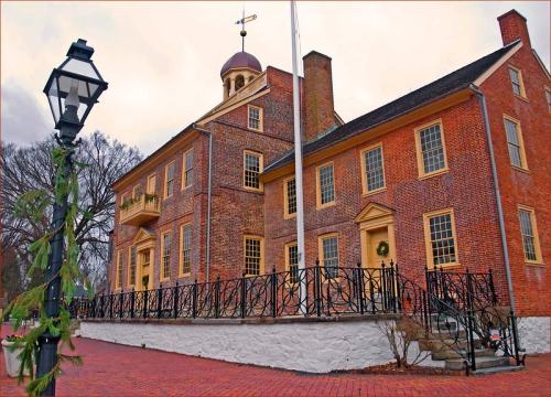 Historic brick building with a cupola, surrounded by a wrought-iron fence and a lamppost, set against a cloudy sky.