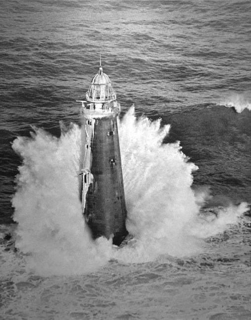 A lighthouse stands tall amidst crashing waves, surrounded by a turbulent sea in black and white.