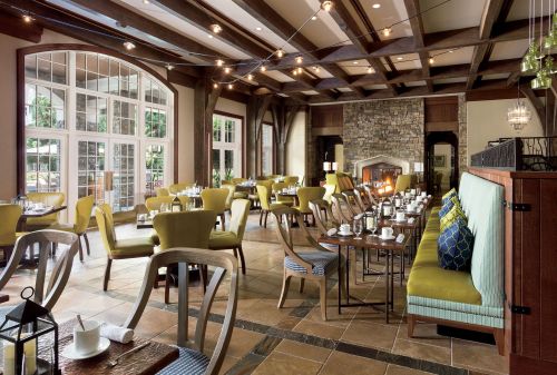 Cozy restaurant interior with wooden beams, green chairs, stone fireplace, and tables set for dining. Warm ambiance.