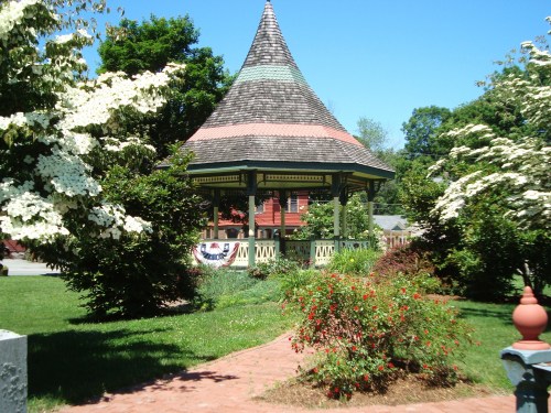 A charming gazebo with a pointed roof surrounded by blooming flowers and greenery on a sunny day.