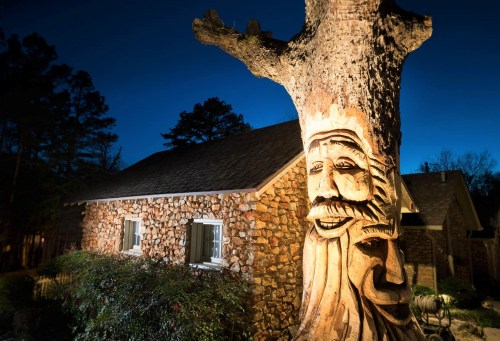 A carved tree trunk with a face stands beside a stone house, illuminated at night with trees in the background.