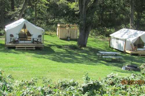 Two canvas tents on wooden platforms in a grassy area, surrounded by trees, with a wooden structure nearby.